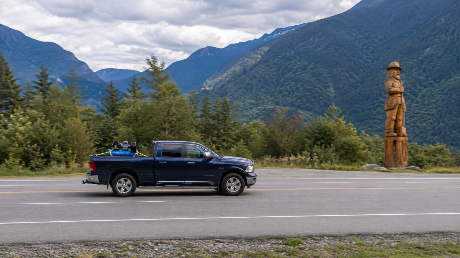 Navy blue delivery truck parked facing a tall wooden chainsaw-carved lumberjack sculpture, with cloudy mountain peaks in the background
