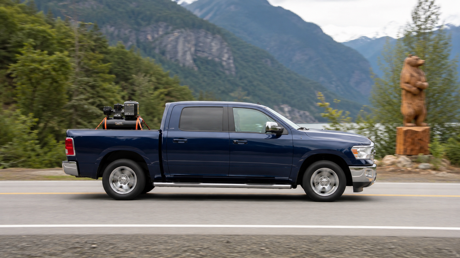 Side profile of the navy blue delivery truck driving past a large wooden bear chainsaw carving on a tree stump, with mountains and forest in the background