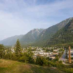 Elevated view over a grassy hillside and pine trees looking down on the town of Hope, BC, in the Fraser Canyon valley, with a tall wooden chainsaw-carved totem in the foreground and forested mountains beyond