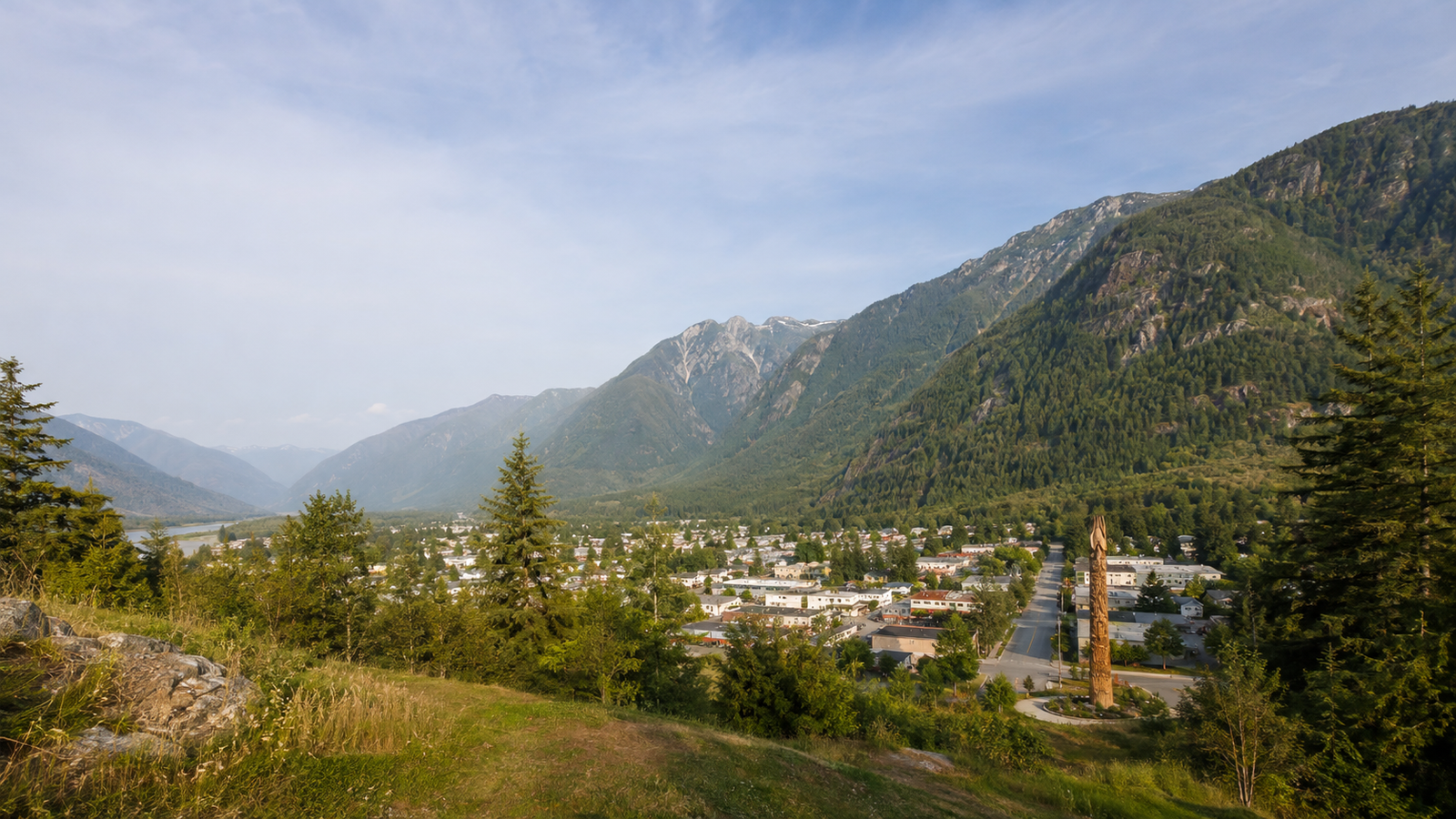 Elevated view over a grassy hillside and pine trees looking down on the town of Hope, BC, in the Fraser Canyon valley, with a tall wooden chainsaw-carved totem in the foreground and forested mountains beyond