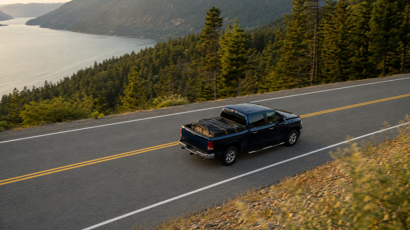 Aerial view of Canyon Runner Hotshot navy blue pickup truck with covered bed, driving along a rural highway near Nelson, BC
