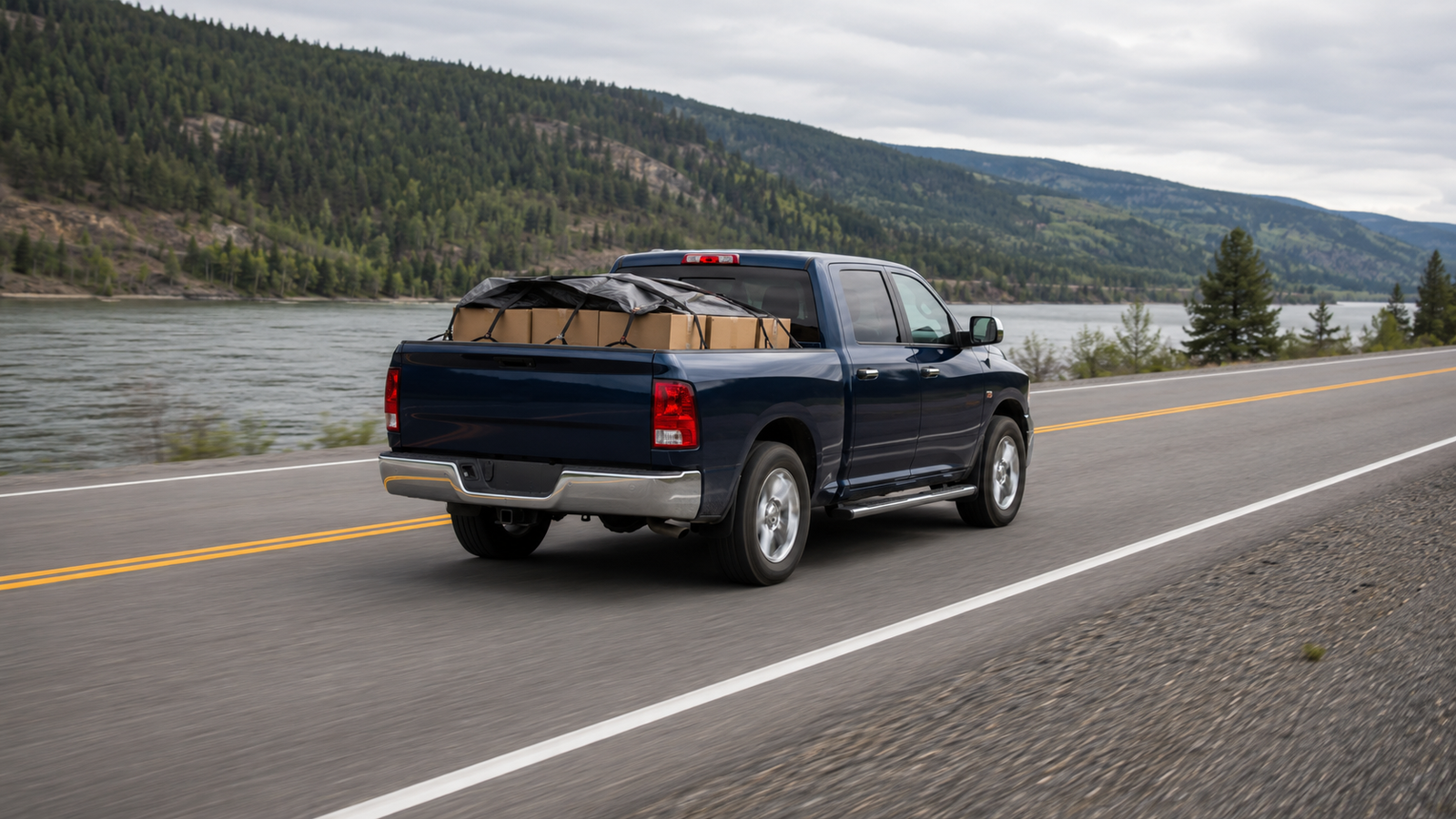 Canyon Runner Hotshot navy blue pickup truck loaded with boxed cargo, driving along a riverside road near Quesnel, BC