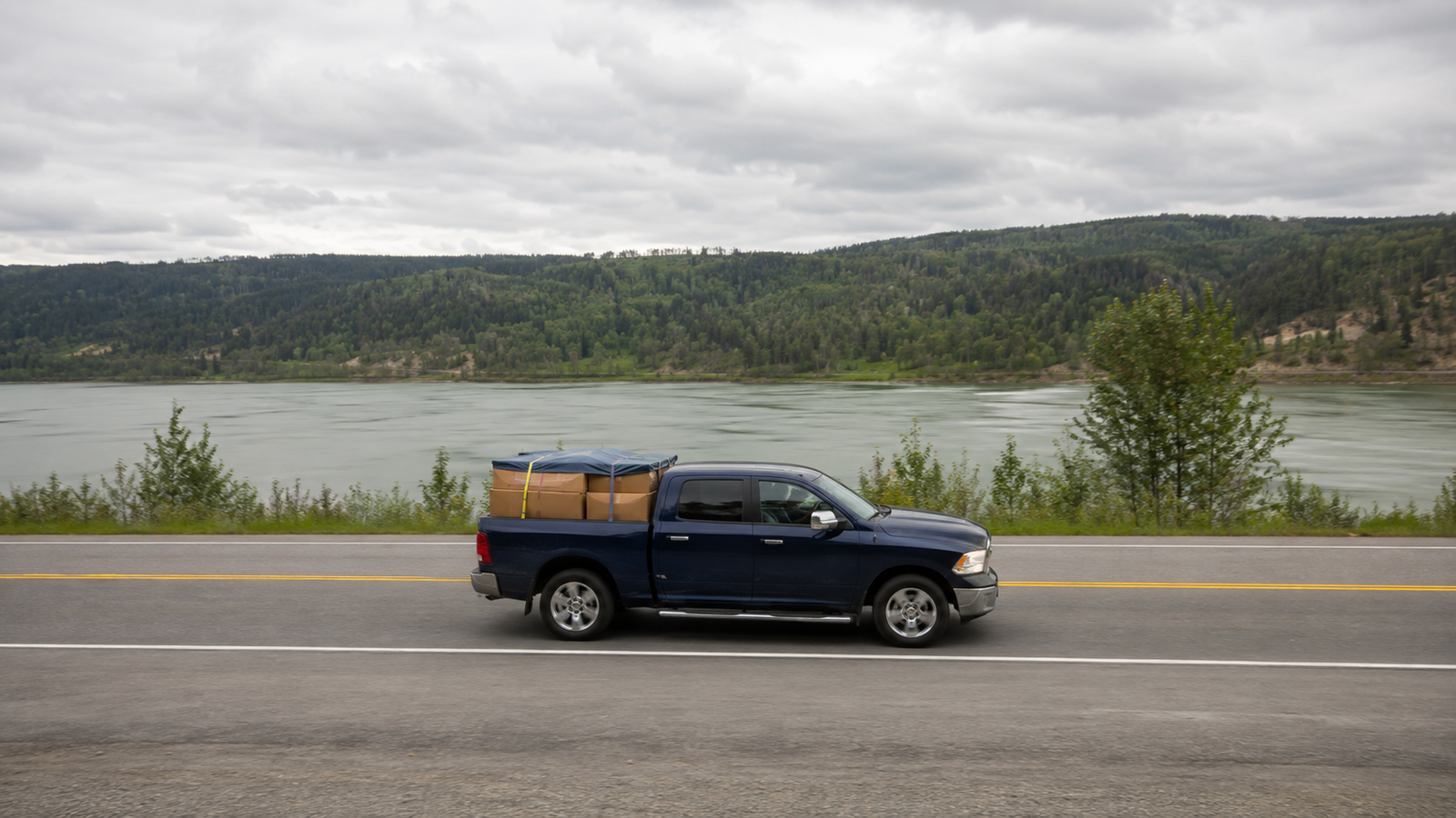 Canyon Runner Hotshot navy blue pickup truck side profile with tarp-covered cargo in the bed, driving beside the Fraser River near Quesnel, BC